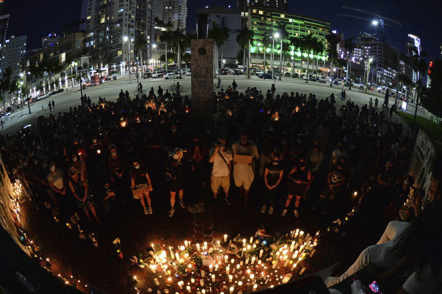 A large group of people are seen standing in front of a candle vigil during a late hour in the night. The only light comes from the multiple candles lit, along with lights from a street and buildings in an area behind the people at the event.