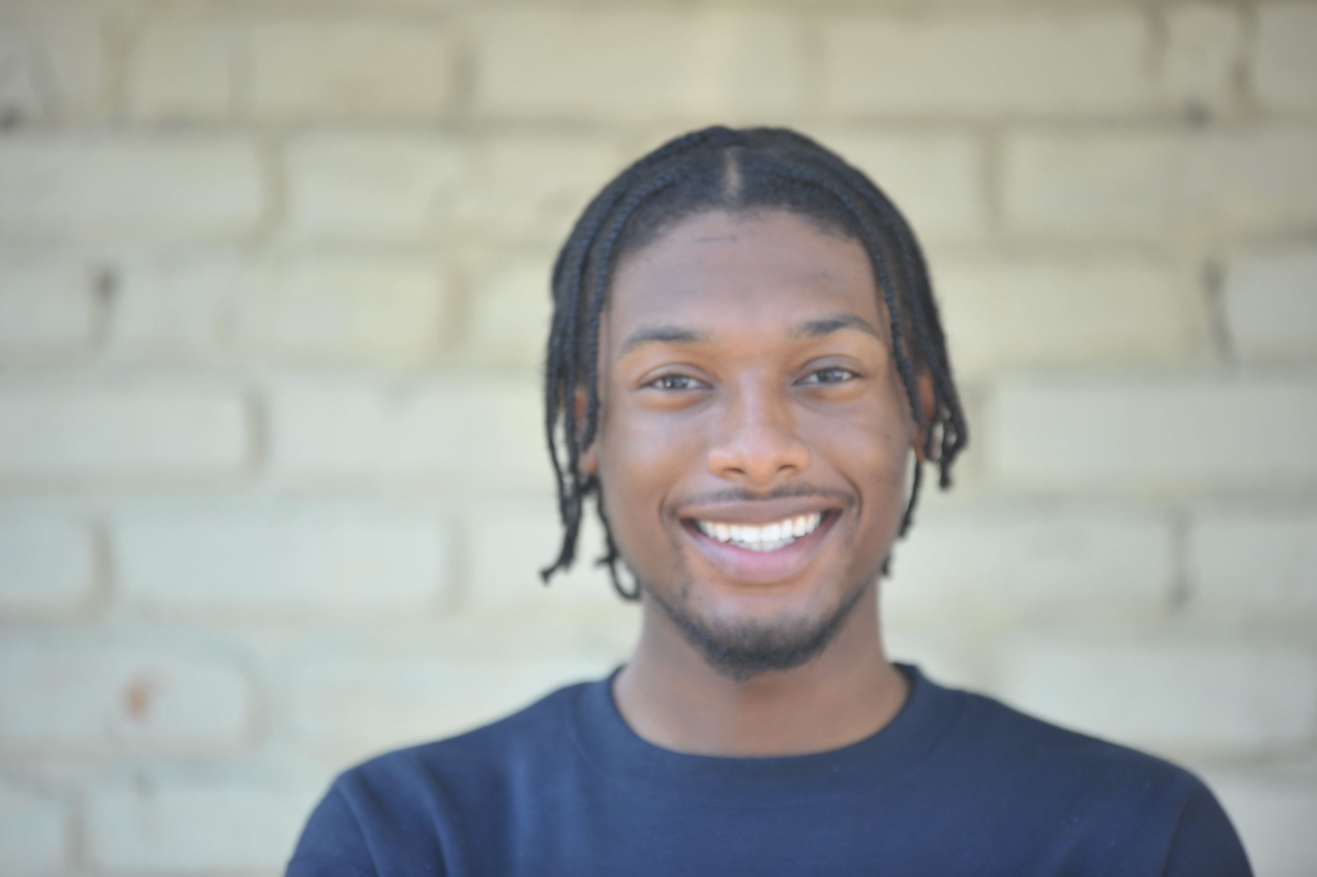 A man with dreadlocks and a goatee stands, on a sunny day, smiling for a photo. He is standing in front of a brick wall. He is wearing a navy blue shirt.