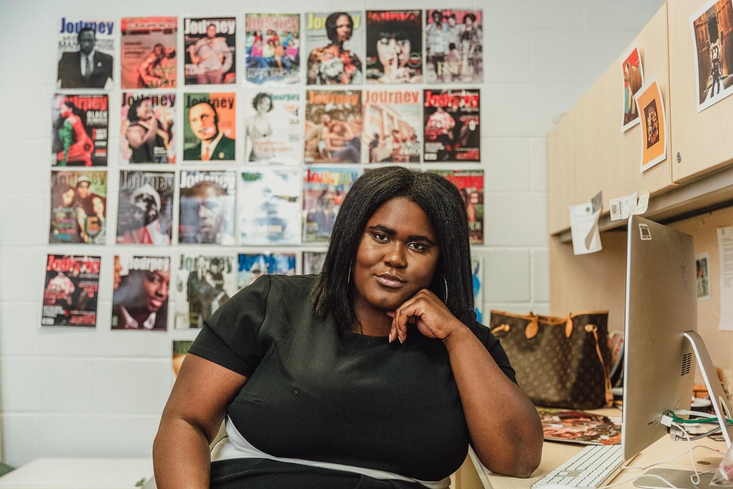 A woman is seen sitting at a desk, posing for a photo with her cheek resting on the knuckle of her hand. In the background, posted on a wall, are magazine covers. A MacBook desktop is seen near the woman, along with a brown purse in the back.