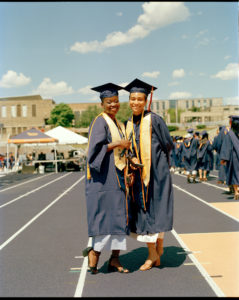 Two women are standing close together, posing for a photo with their navy blue caps and gowns on and yellow-ish gold garments over the caps and gowns. It's a sunny day outside, and the two are smiling. Both are holding a small pole-like object.