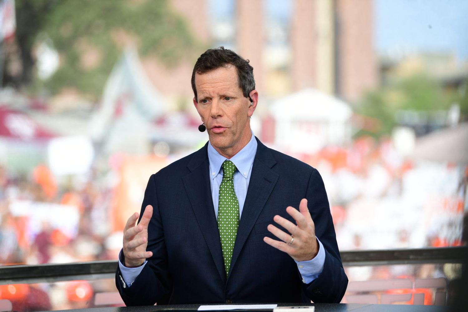 A tall white male with a navy blue suit jacket on and a green tie sits with his hands midway in the air while speaking. It is a sunny day outside, and a crowd is blurred in the background.