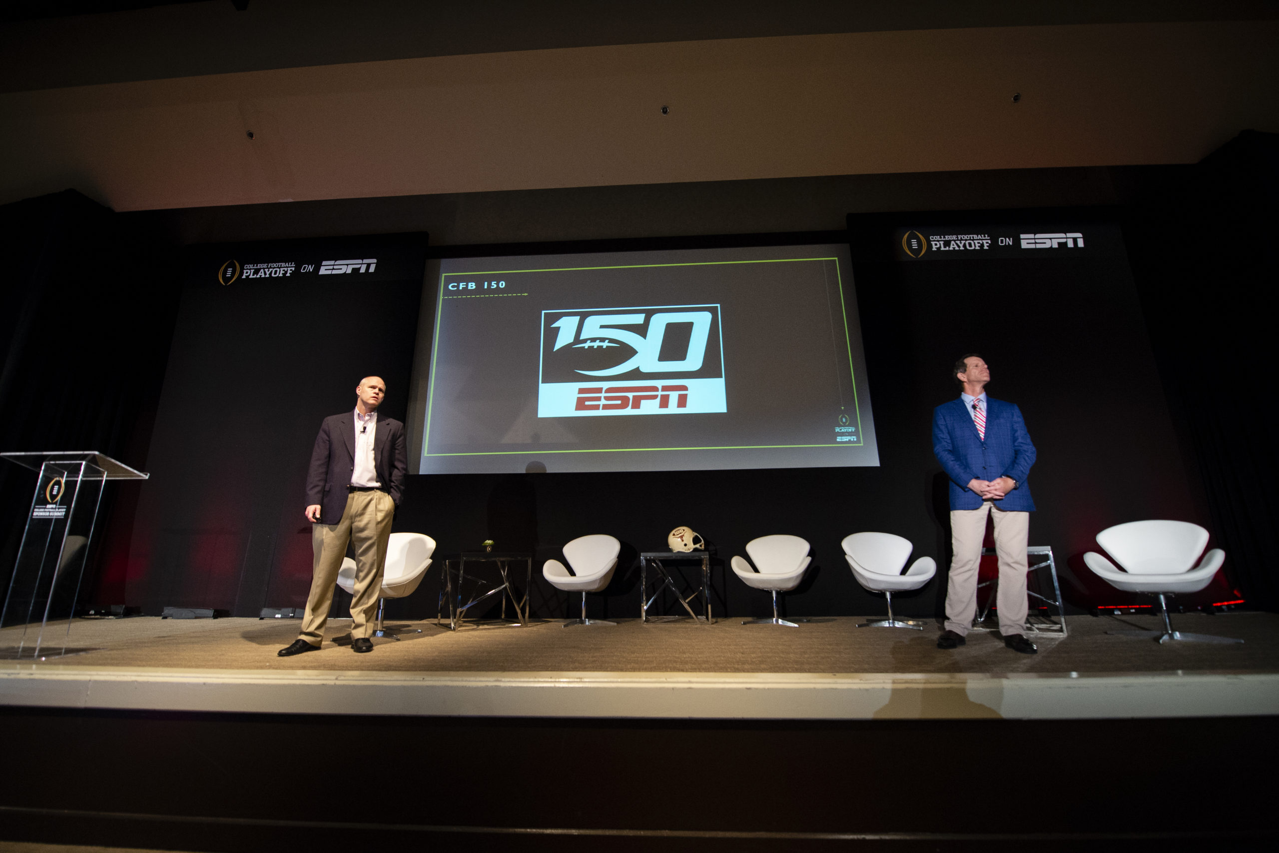 Two males stand on opposite sides of a screen projector, which boasts a "150 ESPN" graphic. The logo represents a project ESPN worked on titled "150." The two men are looking off into a crowd, giving a presentation.