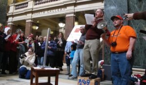 Greg Gordon speaks into a megaphone at the beginning of the airing of grievances, most of them directed at then-Gov. Scott Walker, during a Festivus celebration on Monday, Dec. 23, 2013, in Madison, Wis. (AP Photo/Scott Bauer)