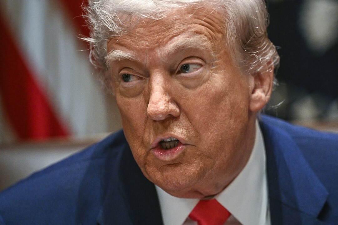 A photo of an white man in his 80s speaking to someone and looking to the left. He's wearing a navy suit and red tie